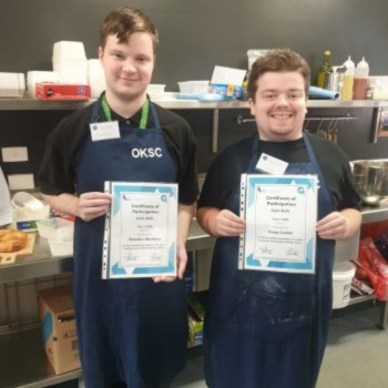 Two young men in aprons smiling and holding certificates of participation in a commercial kitchen.
